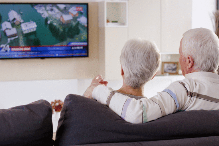 A senior couple sits on a couch watching TV